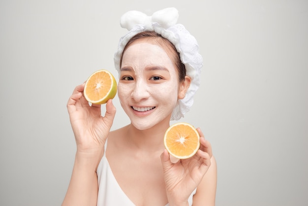 Jovem asiático com máscara de argila facial branca e segurando um pedaço de laranja.