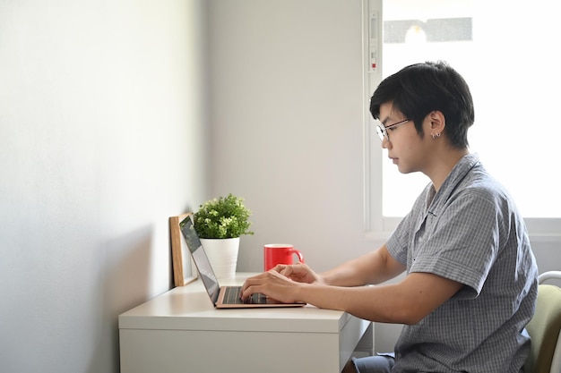 Jovem asiático de pijama, vista lateral, verificando o e-mail no laptop em casa.