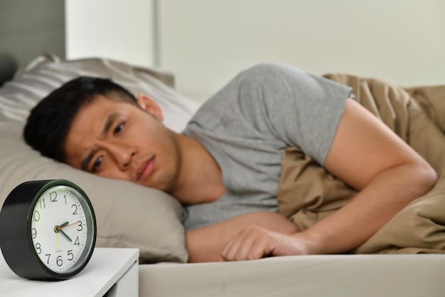 Jovem asiático deprimido deitado na cama não consegue dormir de insônia, concentre-se no despertador