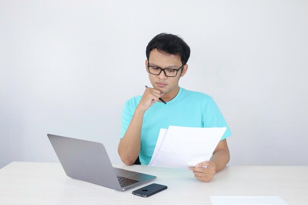 Jovem asiático é sério e se concentra ao trabalhar em um laptop e documento na mesa homem indonésio vestindo camisa azul
