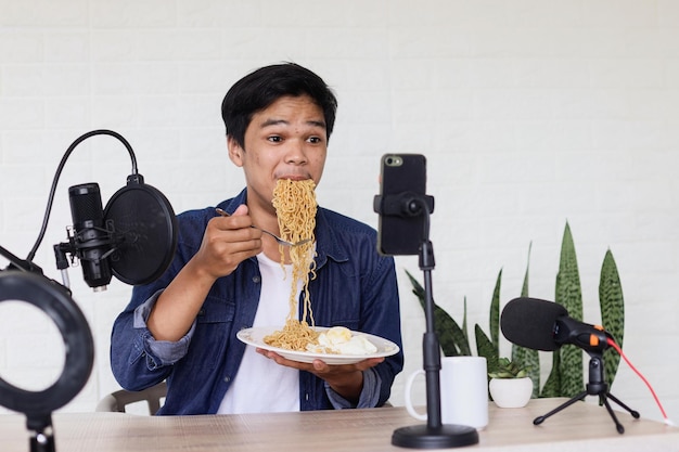 Jovem asiático em estilo casual comendo macarrão na frente do smartphone, gravando vídeo vlog de comida.