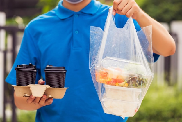 Jovem asiático entregador de uniforme azul usando máscara facial fazendo serviço de mercearia dando caixas de comida de arroz sacos plásticos e café na frente da casa sob pandemia de coronavírus, de volta ao novo conceito normal