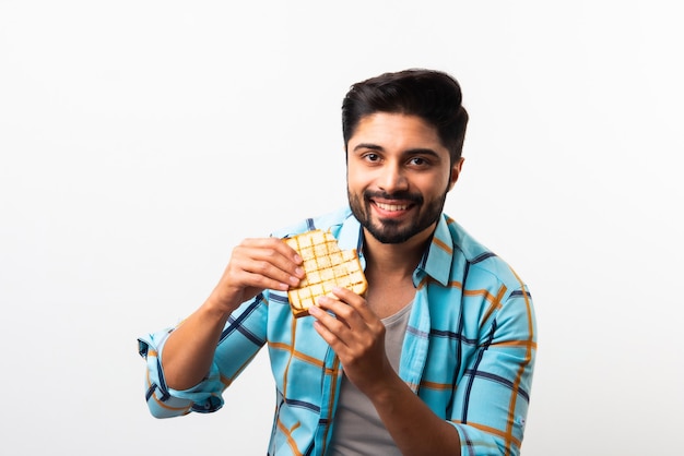 Jovem asiático indiano barbudo come sanduíche de pão enquanto está sentado na cozinha ou na mesa de jantar. Mostrando ou apresentando