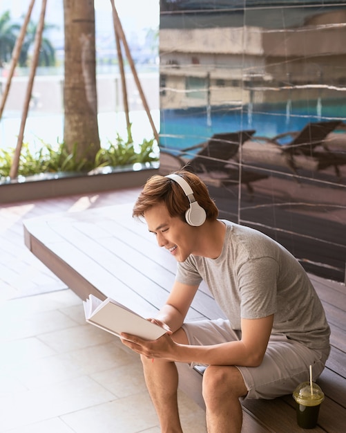 Jovem asiático lendo um livro e ouvindo música à beira da piscina em um dia ensolarado de verão