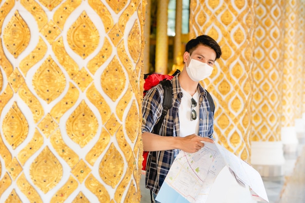 Jovem asiático mochileiro masculino com máscara facial em pé e verificando a direção no mapa de papel na mão no belo templo tailandês.