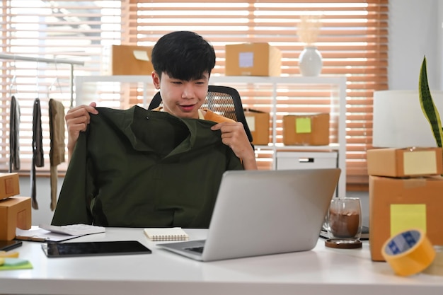 Jovem asiático mostrando roupas na frente do computador portátil enquanto grava vídeo vlog e transmissão ao vivo on-line