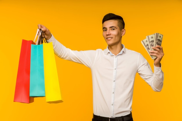 Jovem asiático mostrando seu saco de compras e dinheiro em fundo amarelo