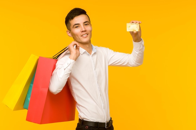 Jovem asiático mostrando sua bolsa de compras e cartão de crédito em fundo amarelo