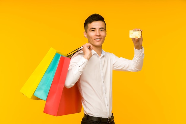 Jovem asiático mostrando sua sacola de compras e cartão de crédito em fundo amarelo
