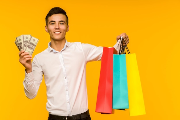 Jovem asiático mostrando sua sacola de compras e dinheiro em fundo amarelo