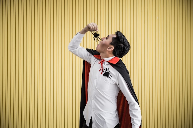 Foto jovem asiático no traje de drácula e comer a aranha preta sobre fundo amarelo do conceito de halloween. retrato de homem adolescente vestido de drácula para comemorar o festival de halloween.