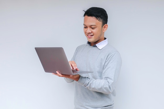 Jovem asiático se sentindo feliz e sorrindo quando está de pé e trabalha laptop indonésio vestindo camisa cinza