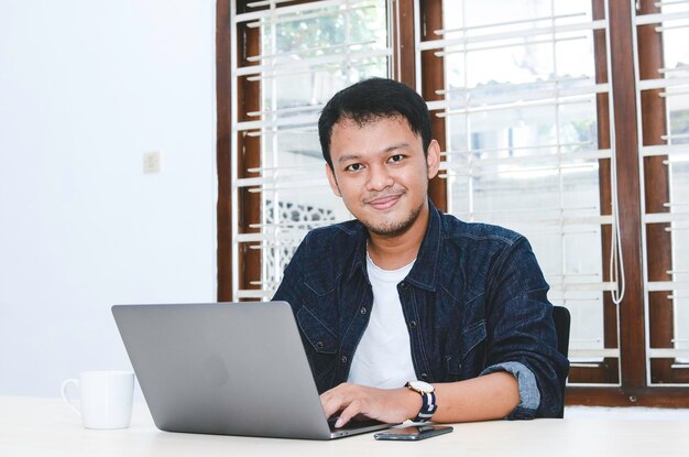 Jovem asiático se sentindo feliz e sorrindo quando trabalha laptop na mesa homem indonésio vestindo camisa azul