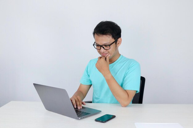 Jovem asiático se sentindo feliz e sorrindo quando trabalha laptop na mesa homem indonésio vestindo camisa azul