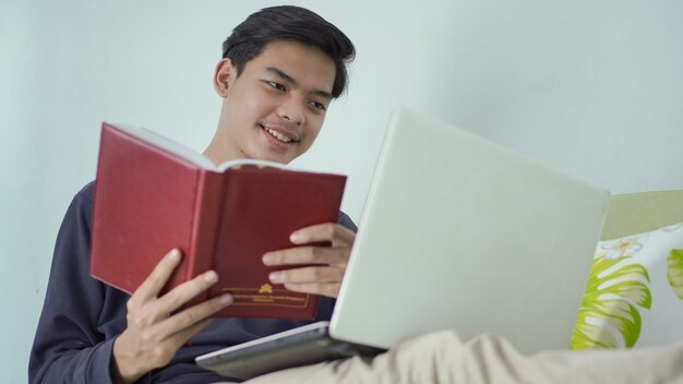 Jovem asiático segurando um livro alegremente enquanto olha para a tela do laptop em casa