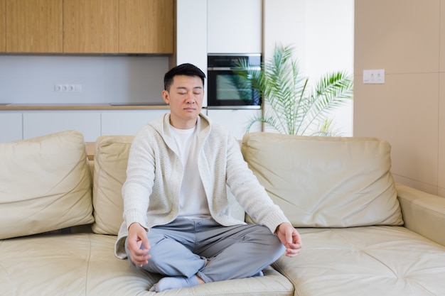 Jovem asiático sentado no sofá em posição de lótus e meditando na calça