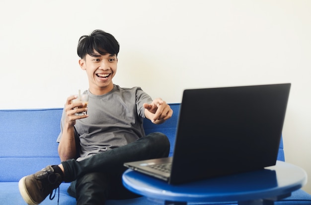 Jovem asiático sentado no sofá tomando café enquanto assiste a um filme em um laptop