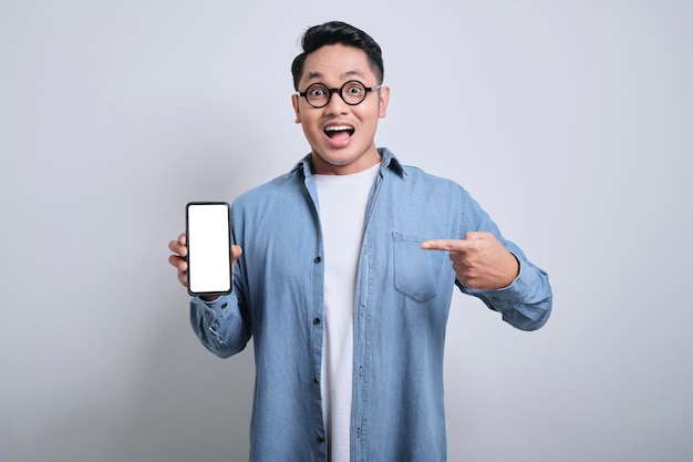 Jovem asiático sorridente vestindo camisa jeans apontando para o celular enquanto mostra a tela vazia em branco