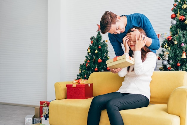 Jovem asiático surpreende e fecha os olhos com um presente de Natal em casa com uma árvore de Natal ao fundo Imagem com espaço de cópia