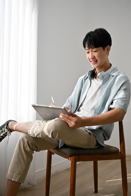 Jovem asiático usando tablet projetando sua obra de arte em tablet sentado em uma cadeira na sala de estar