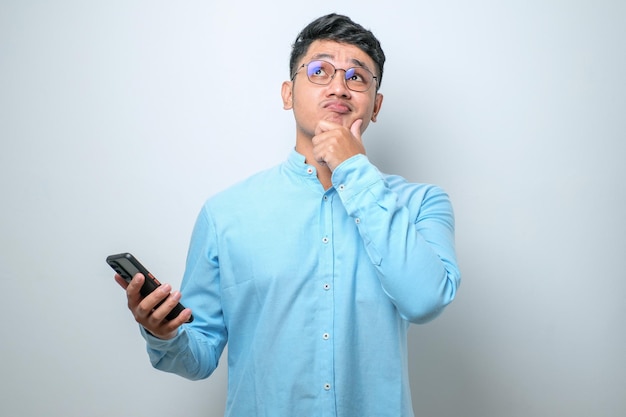 Jovem asiático usando telefone celular pensando e olhando para cima enquanto a mão no queixo