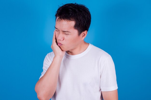 Jovem asiático veste uma camiseta branca com dentes sensíveis ou dor de dente sobre fundo azul. conceito de saúde.
