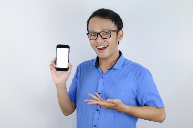 Jovem asiático vestindo camisa azul está de pé e sorrindo apontando para o espaço em branco na tela do smartphone em fundo branco