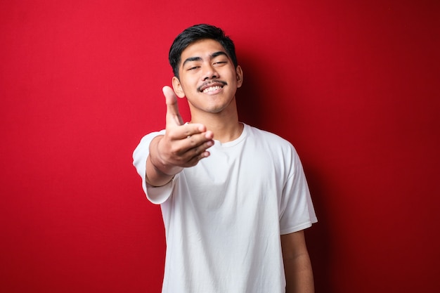 Jovem asiático vestindo camiseta casual sorrindo amigável oferecendo um aperto de mão como saudação e boas-vindas sobre fundo vermelho. negócio de sucesso.