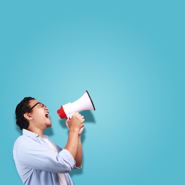 Jovem asiático vestindo gritando com megafone sobre fundo azul com espaço de cópia