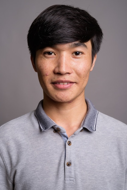 Jovem asiático vestindo uma camisa pólo cinza contra uma parede cinza