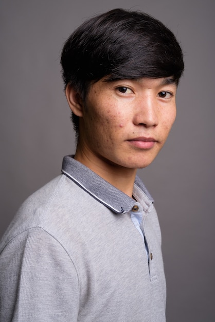 Jovem asiático vestindo uma camisa pólo cinza contra uma parede cinza