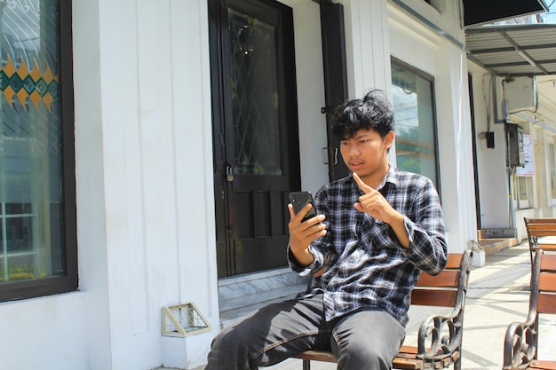 jovem asiático zangado apontando e ameaçando quando faz uma videochamada usando seu smartphone e sentado