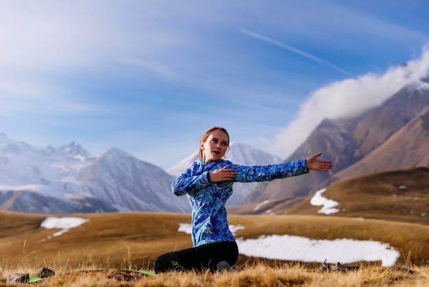 Jovem ativa, com um casaco azul, pratica ioga no contexto das montanhas do Cáucaso, viaja