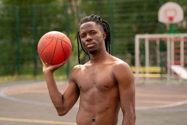 Jovem atleta africano musculoso sem camisa com bola em pé na quadra de basquete