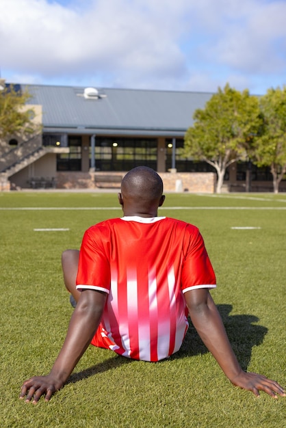 Jovem atleta afro-americano sentado na grama no campo ao ar livre voltado para fora