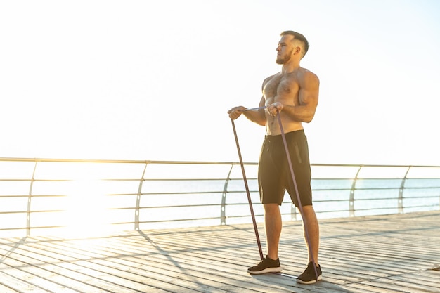 Jovem atleta com o tronco nu se exercita com bandas de borracha de fitness no início da manhã na praia Treinamento muscular Estilo de vida saudável