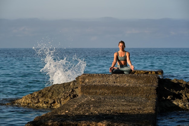 Jovem atleta feminina fazendo pose de Padmasana com as mãos em gyan mudra enquanto está sentada em uma pedra e praticando ioga à beira-mar