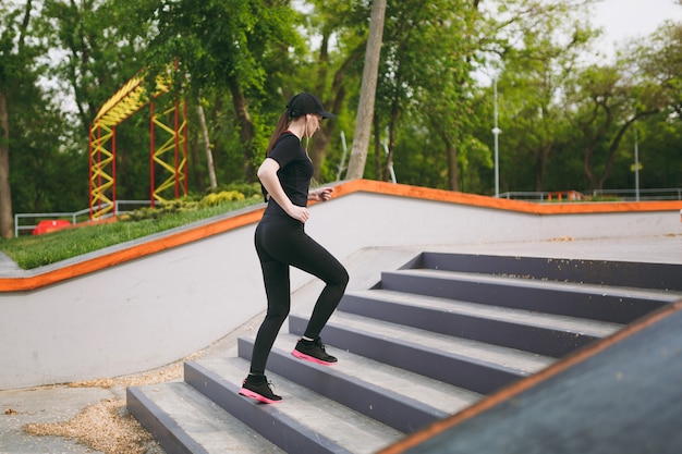 Jovem atlética linda morena de uniforme preto, boné com fones de ouvido, fazendo exercícios de esporte, treinando e correndo, subindo em escadas no parque da cidade ao ar livre