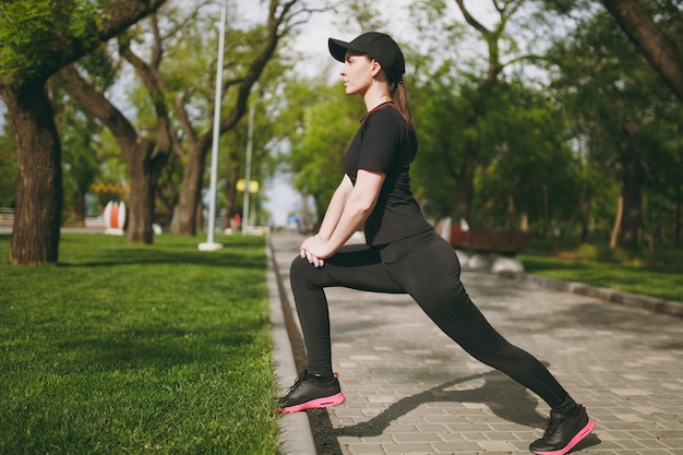 Jovem atlética linda morena de uniforme preto boné fazendo exercícios de alongamento, aquecimento antes de correr ou treinar, parado no parque da cidade ao ar livre