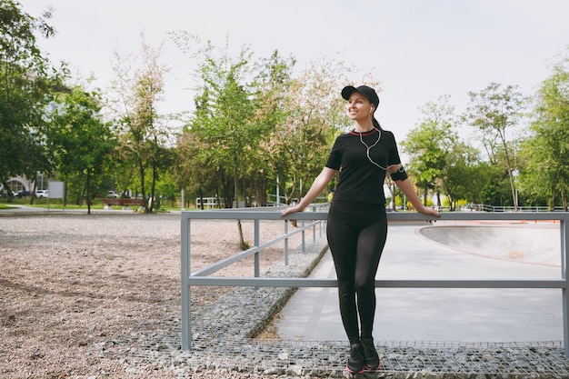 Jovem atlética linda morena de uniforme preto e boné com fones de ouvido, ouvindo música, antes ou depois de correr, treinando no parque da cidade ao ar livre