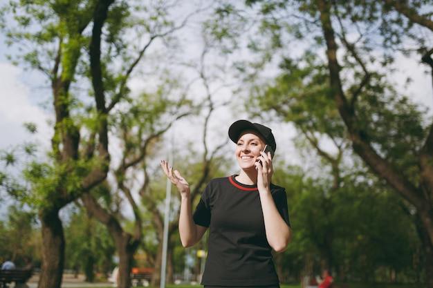 Jovem atlética sorridente linda morena de uniforme preto e boné falando no celular durante o treinamento, olhando para o lado e parada no parque da cidade ao ar livre