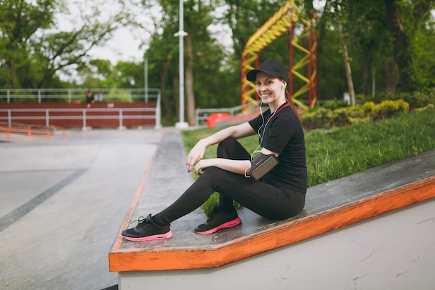 Jovem atlética sorridente linda mulher de uniforme preto e boné com fones de ouvido, ouvindo música, descansando e sentado antes ou depois de correr, treinando no parque da cidade ao ar livre