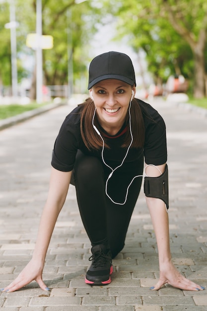 Jovem atlética sorridente morena de uniforme preto e boné com fones de ouvido, ouvindo música no início baixo antes de correr, treinando no caminho no parque da cidade ao ar livre