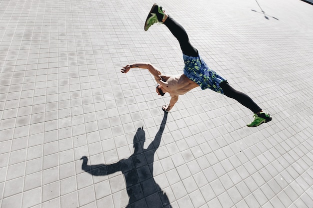 Jovem atlético com um torso nu com tatuagens vestida com legging preta e shorts azuis fazendo o truque, de pé por um lado nas lajes na rua.