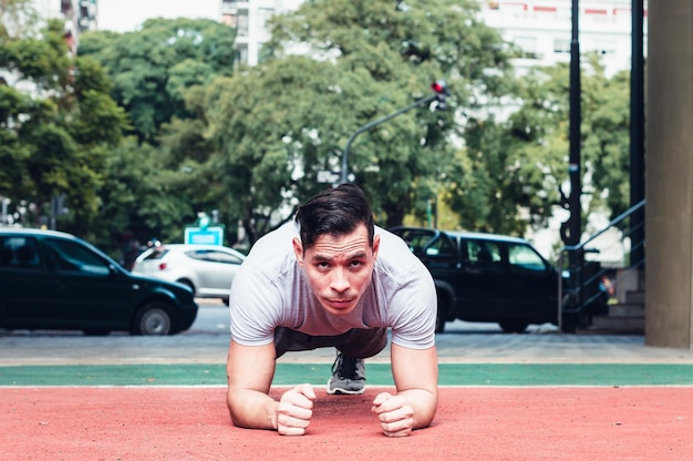 Foto jovem atlético latino, pele branca, fazendo flexões, vestido com roupas cinza e em uma praça de fitness com ambiente urbano, ao ar livre.