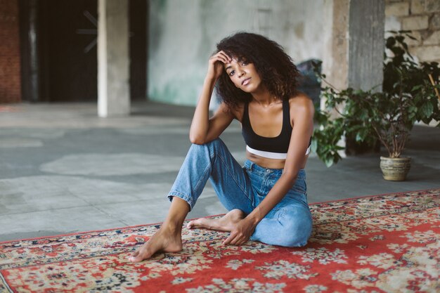 Jovem atraente afro-americana com cabelo escuro encaracolado, vestindo um top esportivo e jeans, pensativa, enquanto está sentada no tapete vintage em casa