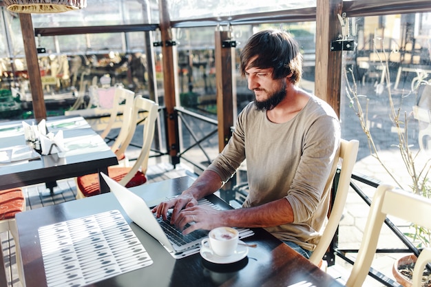 Foto jovem atraente barbudo digitando no laptop no café ao ar livre