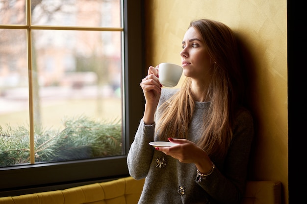 Jovem atraente, bebendo café ou chá no café e olhando pela janela.