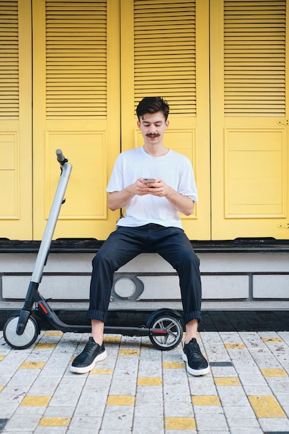 Jovem atraente bigode sentado com e-scooter enquanto sonhadoramente usando celular com fundo amarelo na rua da cidade