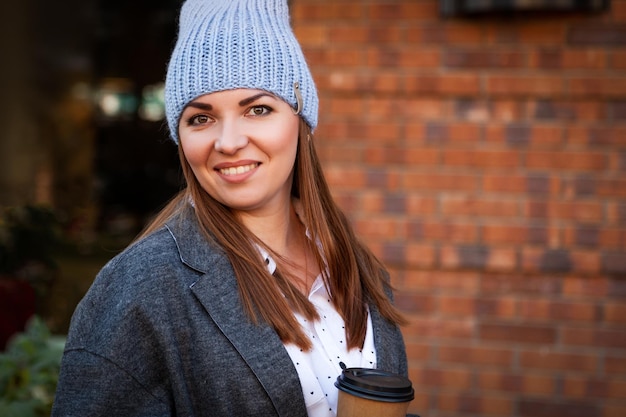 Jovem atraente de chapéu de tricô cinza e casaco cinza bebe café para ir Sorrindo linda mulher feliz hipster na rua da cidade vestindo roupas elegantes Estilo de outono urbano stydent